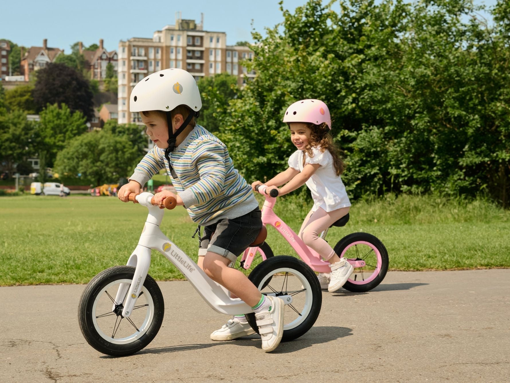 wo young children riding LittleLife balance bikes in a park, wearing safety helmets