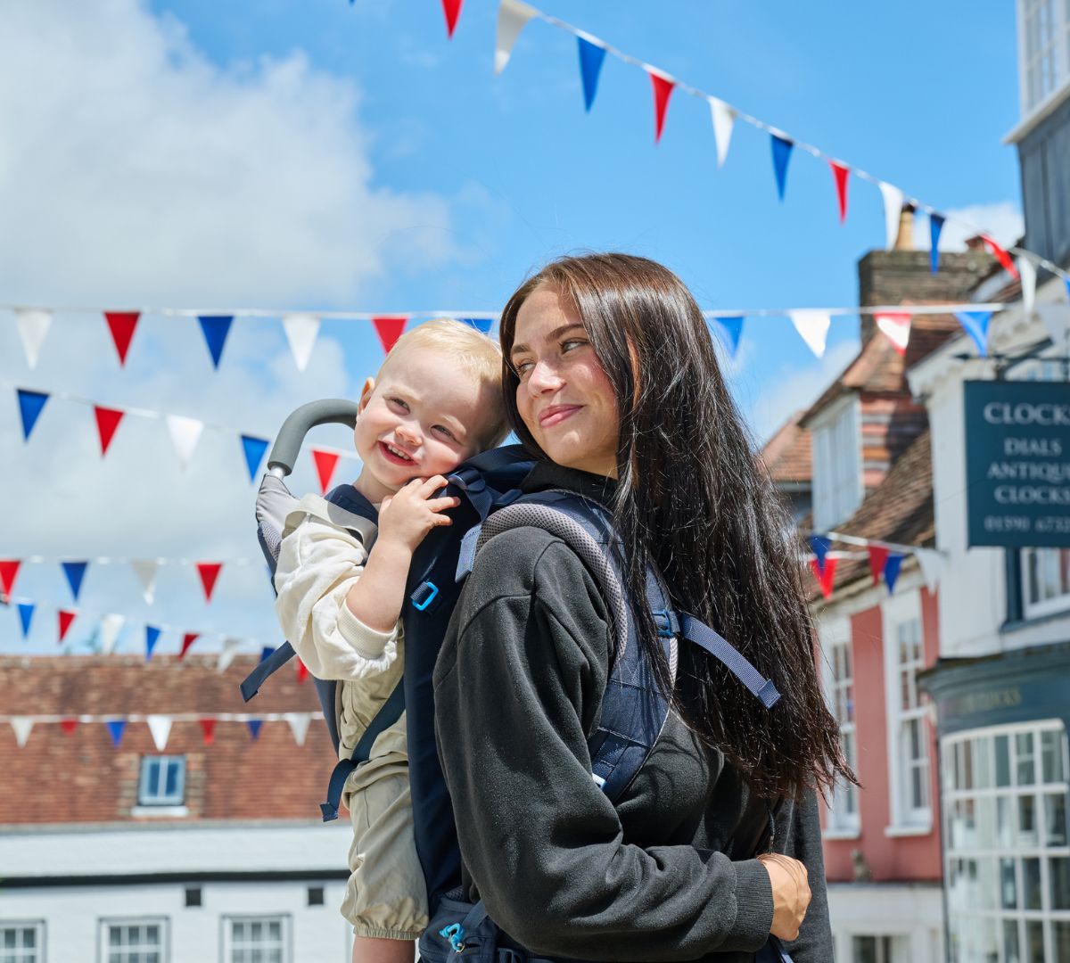 Woman smiling as she carries a happy toddler in a LittleLife child carrier, with colourful bunting in the background.