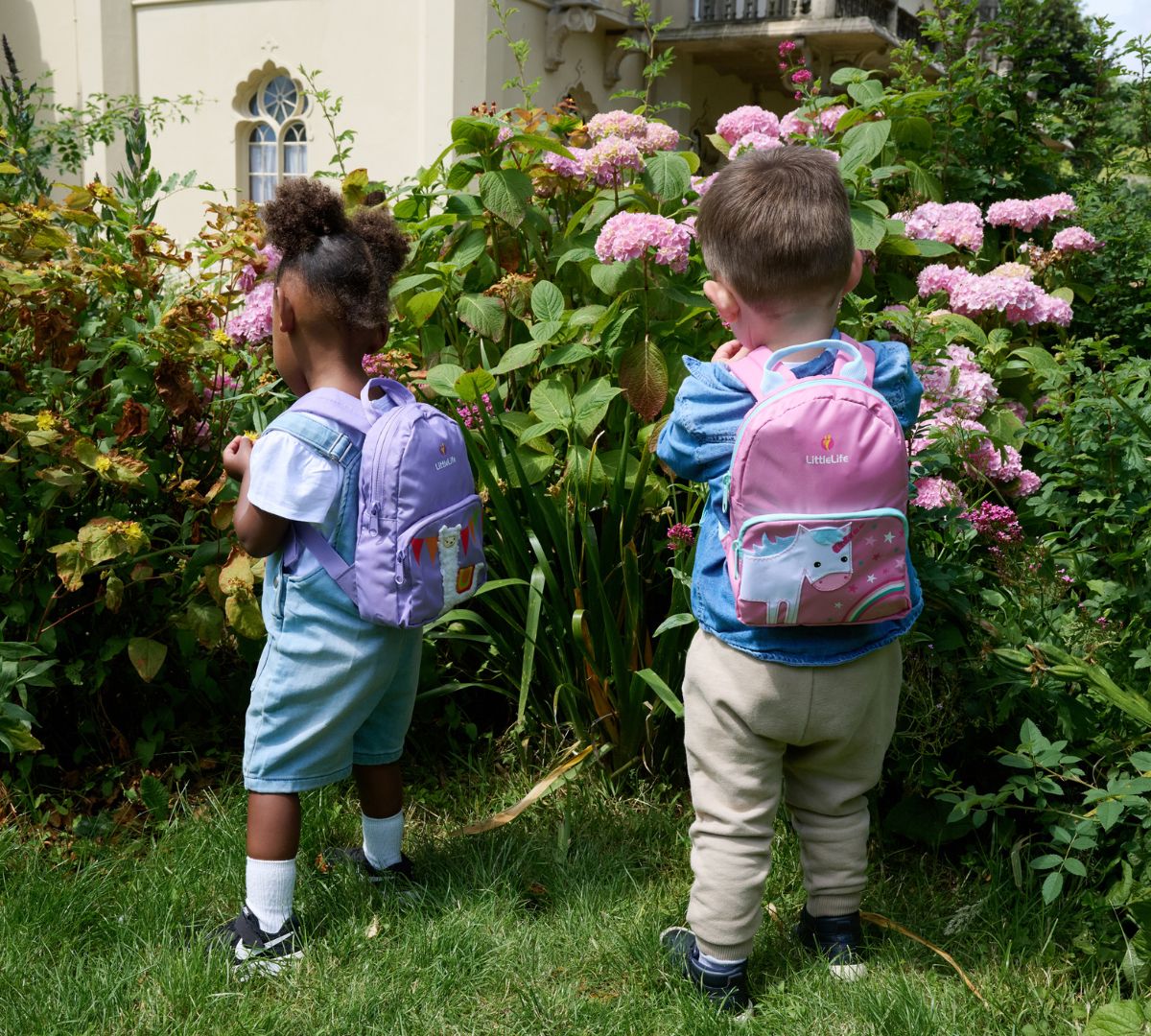 Two young children exploring a garden, each wearing a LittleLife toddler backpack in fun, pastel designs.