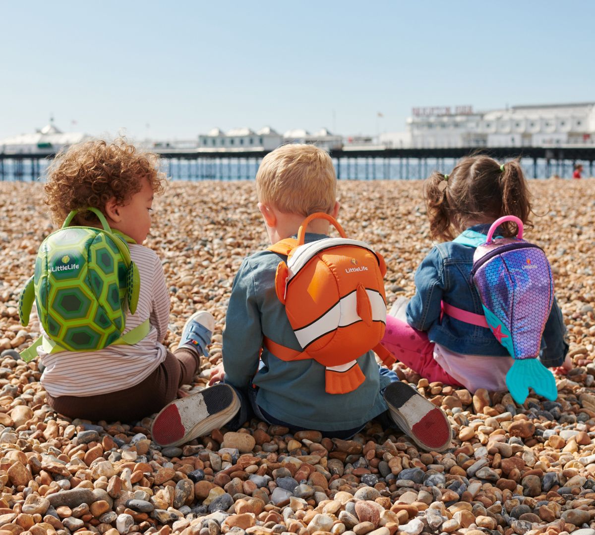 Three children sitting on a pebble beach, each wearing a sea creature-themed LittleLife backpack including a turtle, clownfish and mermaid tail design.